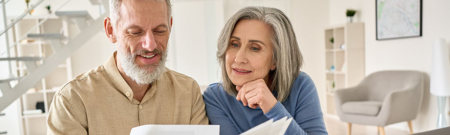 Middle aged couple reading paper bills calculating pension using laptop at home.