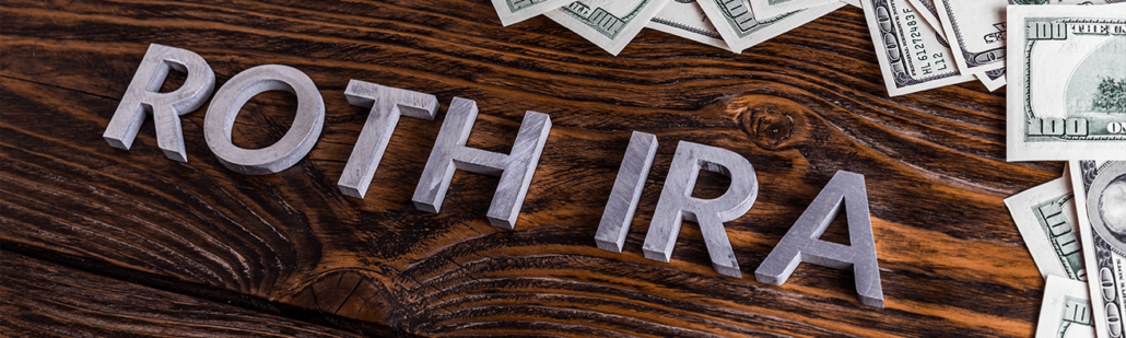 The words Roth IRA in metallic silver lying on a wooden table with hundred dollar bills lying to the top and right side