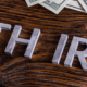 The words Roth IRA in metallic silver lying on a wooden table with hundred dollar bills lying to the top and right side