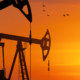Two oil derricks are silhouetted against an orange and yellow sunset
