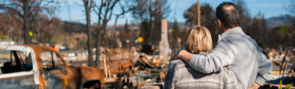 A man drapes his arm over a woman's shoulder as they face away from the camera. It appears they are looking at damage to a house and vehicle caused by a fire.