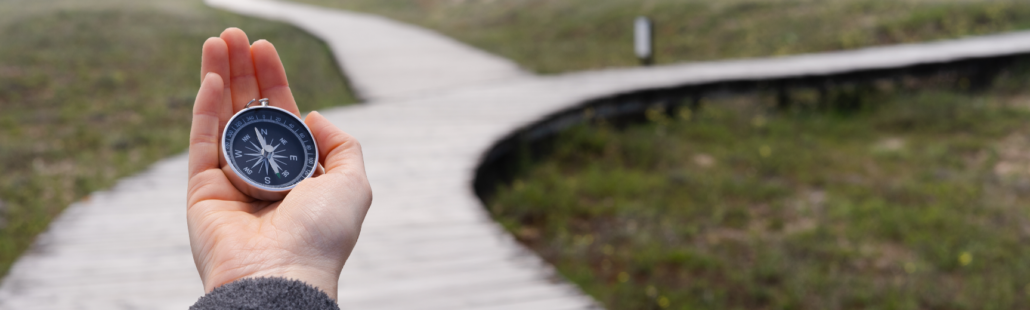 A hand holds a compass against the background of a wood plank path that eventually diverges into two paths.